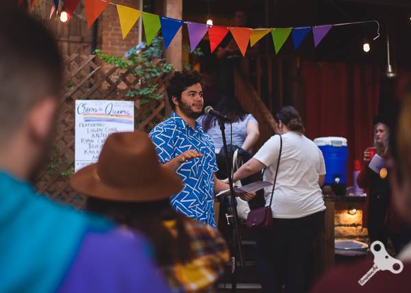 James stands at a microphone outside at Wine Squared wearing a blue button down shirt and under rainbow pennants.