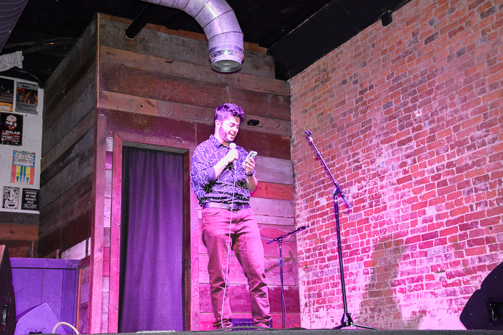James stands at a microphone on a raised stage with a brick wall next to him. He's wearing a long-sleeve, black button down and red pants.