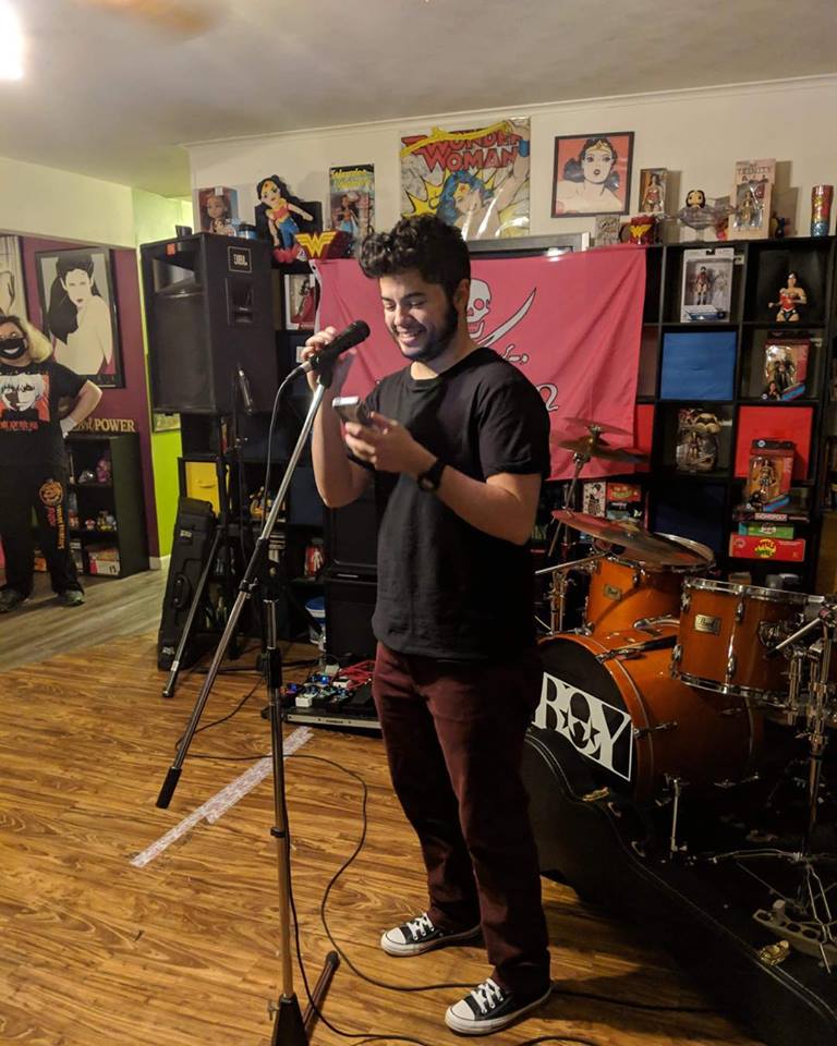 James stands at a microphone in front of a drumset in a house. He's wearing a black short-sleeved shirt, red pants, and black converse high-tops.