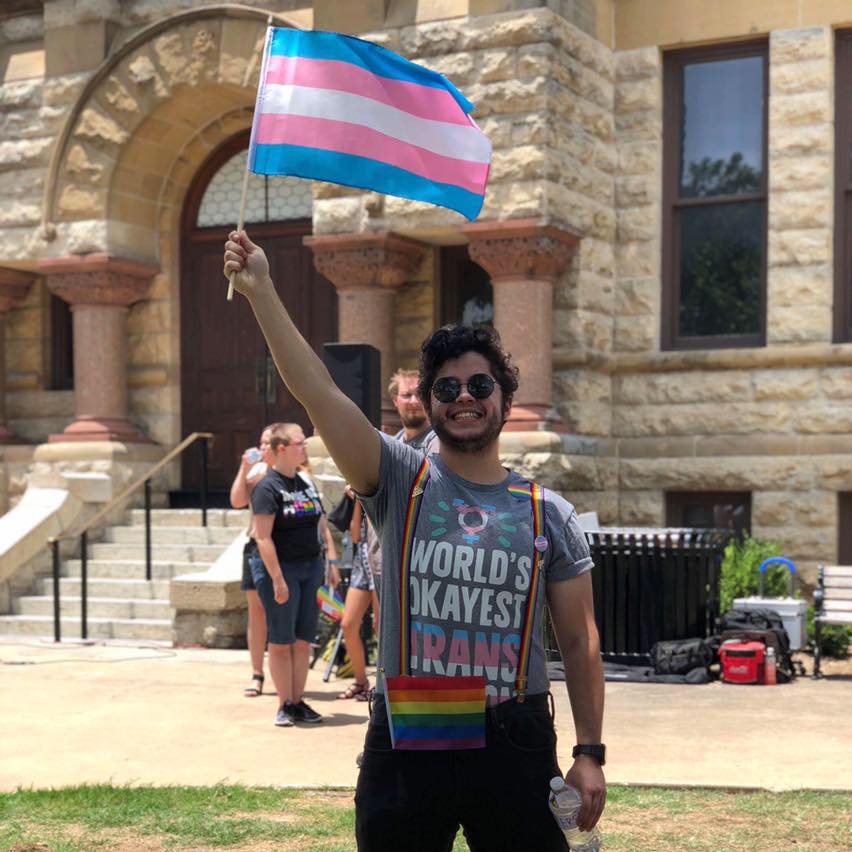 Photo of James in front of the Denton County Courthouse. He is grinning and wearing a shirt that reads: Word's Okayest Trans Person" as he waves a trans flag high above his head.