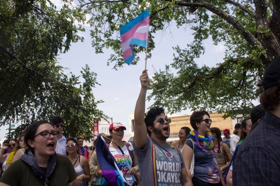 Photo of a Pride march with James in the foreground shouting and holding up a trans flag.