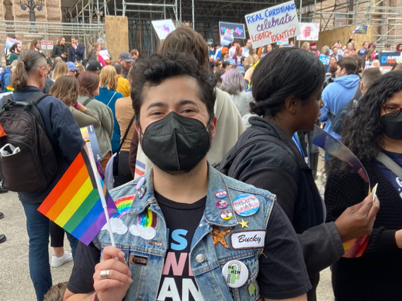 James stands in front of the state capitol in a denim vest with various LGBTQIA+ themed pins. He's wearing a mask and holding up a Pride flag.